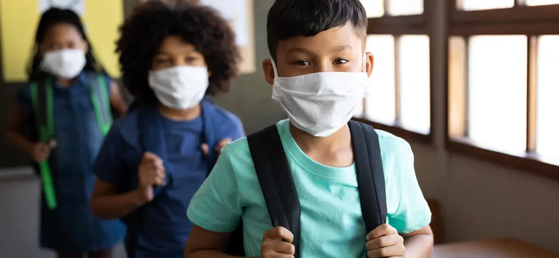 Children wearing face masks and backpacks walking down a school hallway towards the camera.