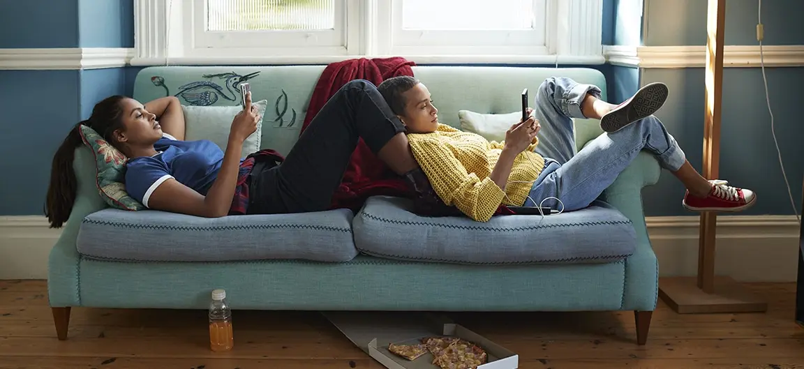 Two teenagers relaxing on a sofa and playing on their phones. They are lying sideways on the cushions of the sofa and are resting on each other and the arms of the chair.