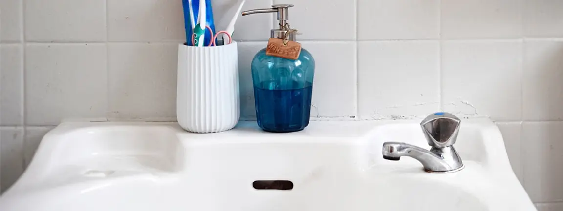 A clean sink in a bathroom. A pot containing toothbrushes and a liquid soap dispenser are on the rim of the sink behind  the faucet.