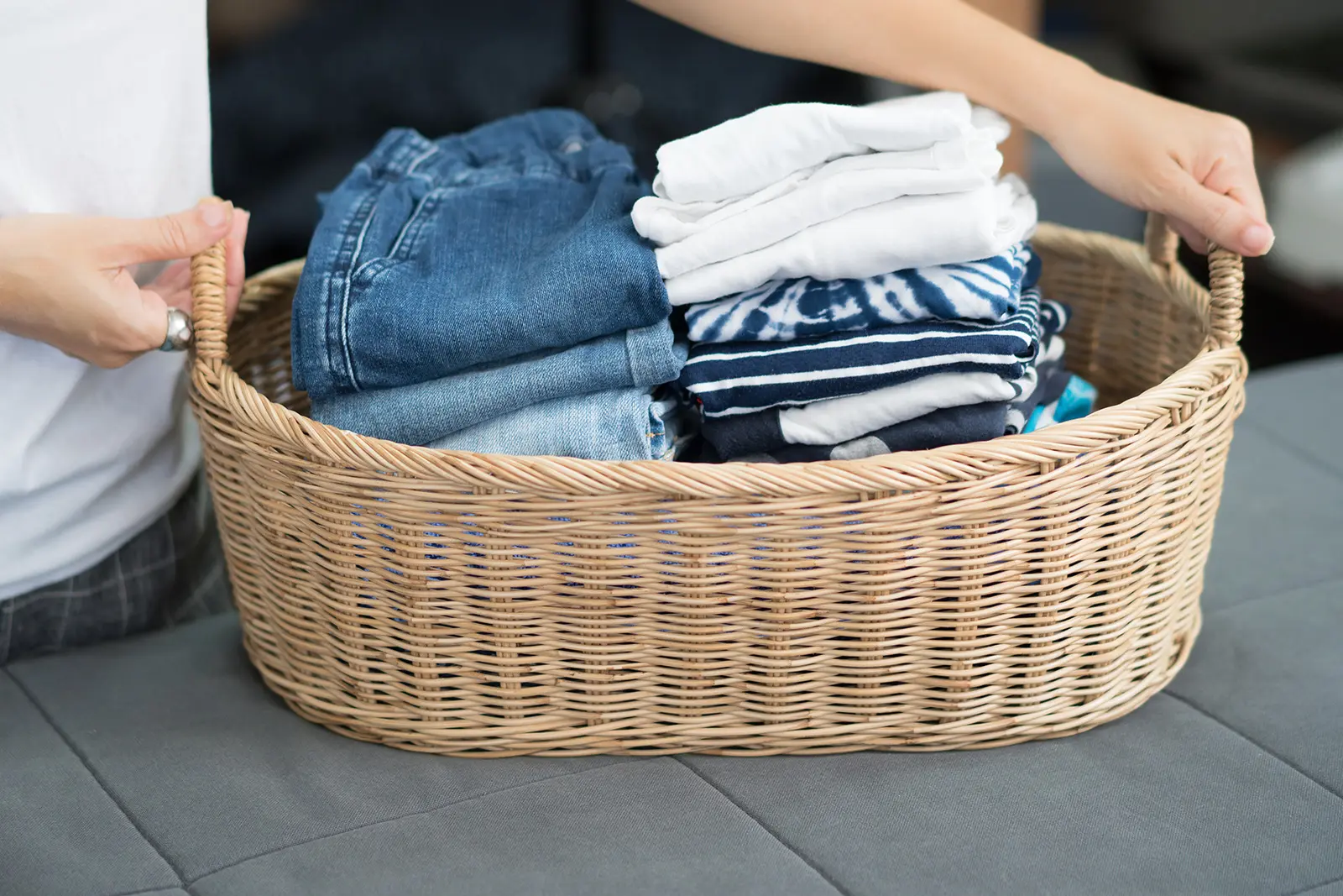 Person holding handles of straw laundry basket with neatly stacked clothes inside