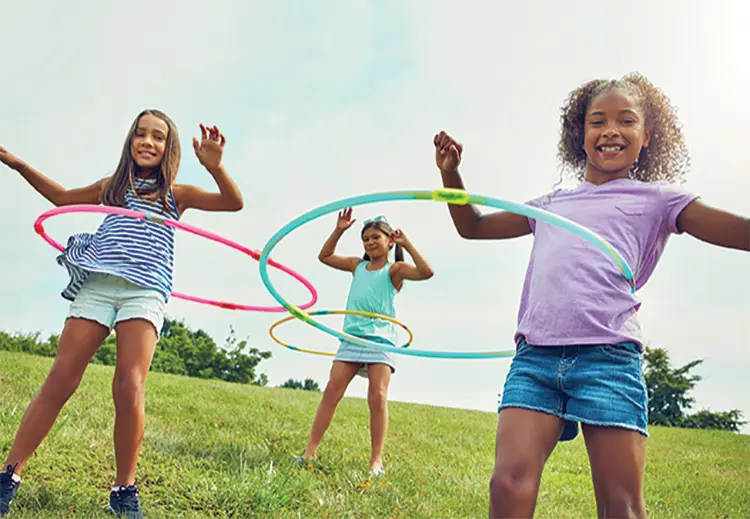 Three children stand in grass field spinning colorful hula hoops on their waists