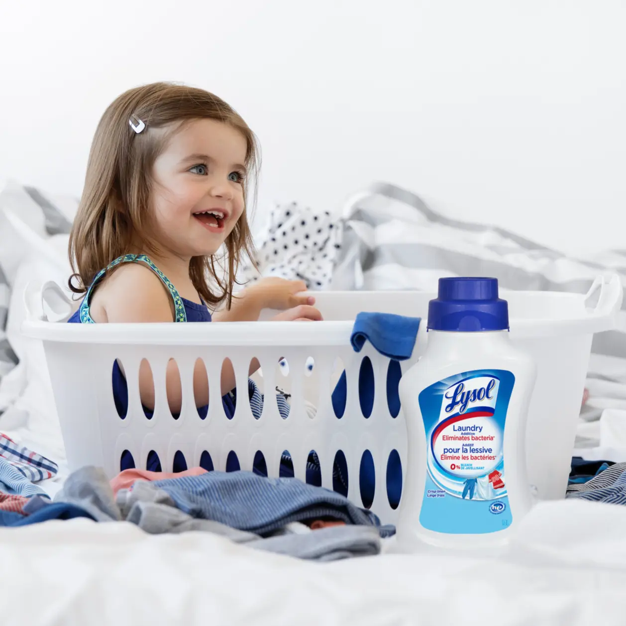 Young child sitting in plastic laundry basket and smiling surrounded by laundry and container of Lysol Laundry Sanitizer