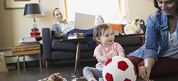 Parent and child in foreground sit on floor with stuffed ball while another parent sits with laptop on sofa in background