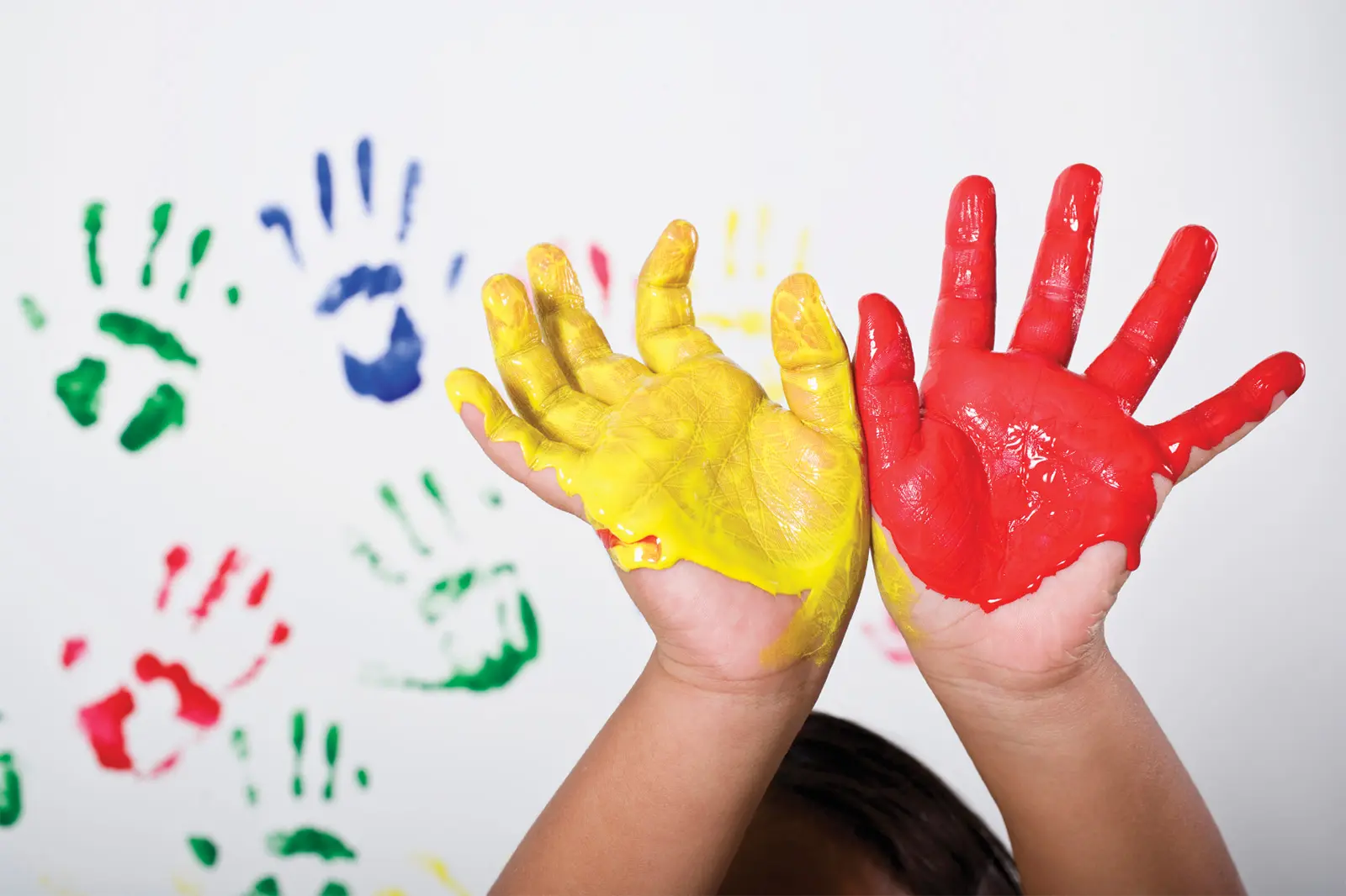 Hands of child covered in paint in front of white surface with paint handprints