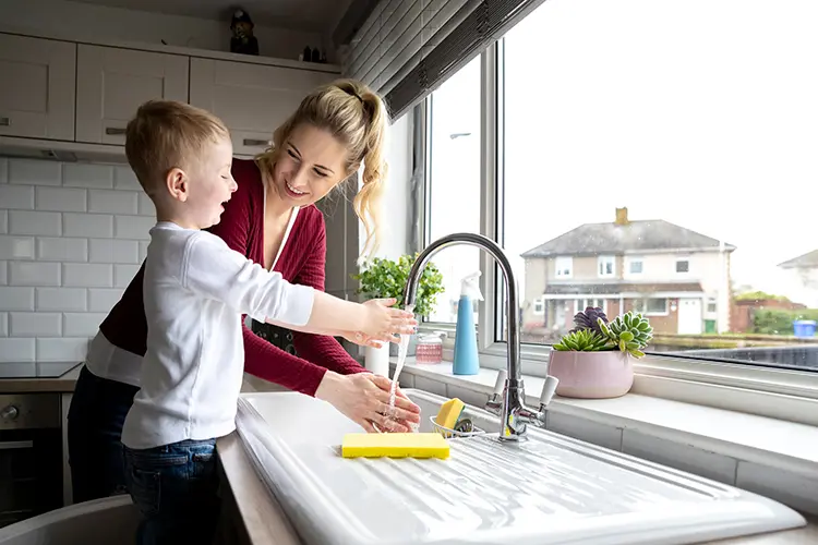 Parent leaning over kitchen sink next to child standing on chair holding hands under running faucet