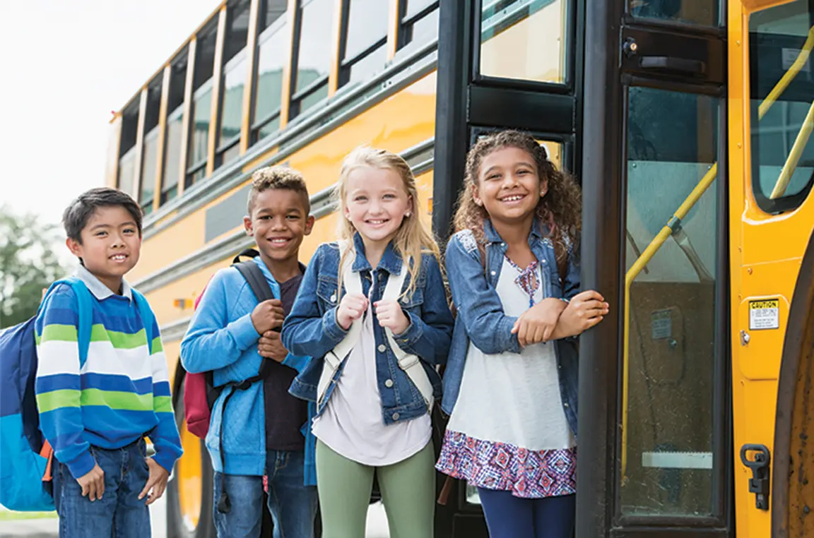 3 children with jackets and backpacks smiling in front of school bus door