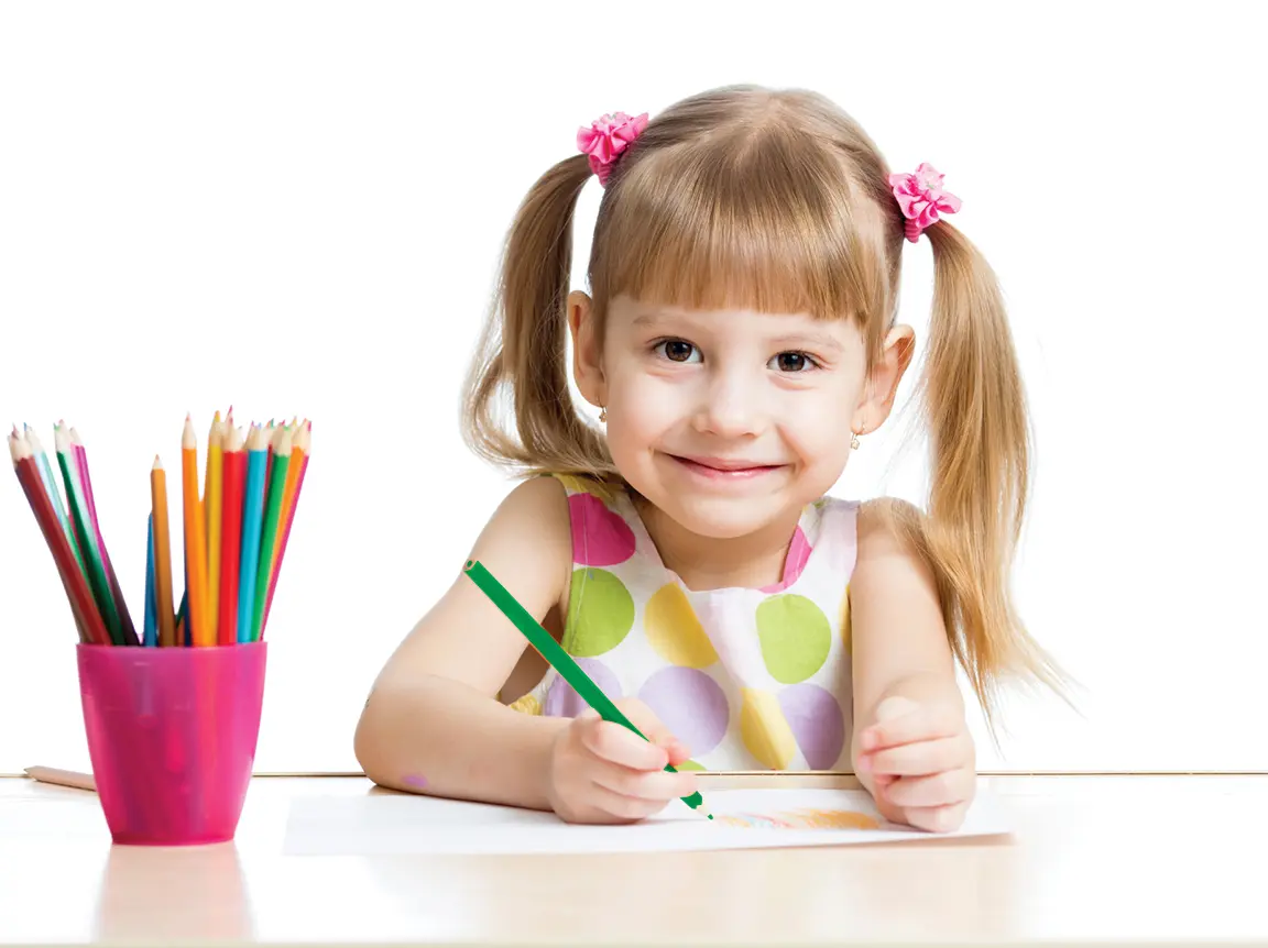 Child next to pot of pencils smiling at camera and drawing on paper