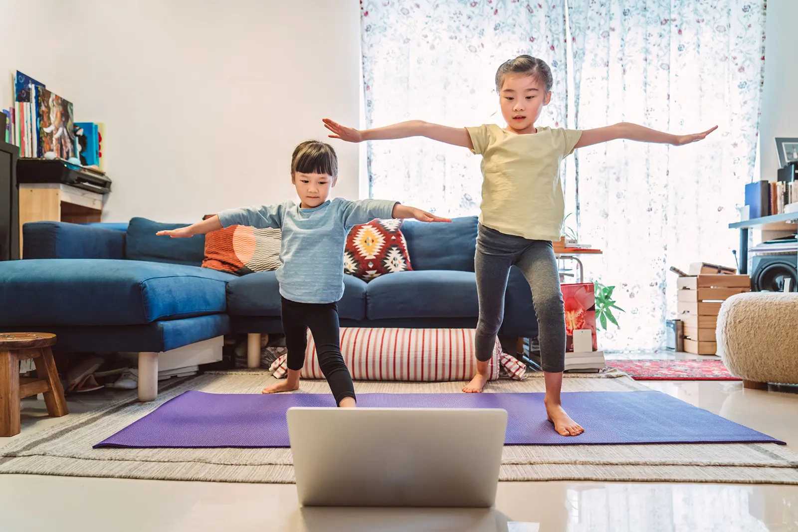 Two children doing yoga pose while standing on yoga mat in living room and looking at laptop screen