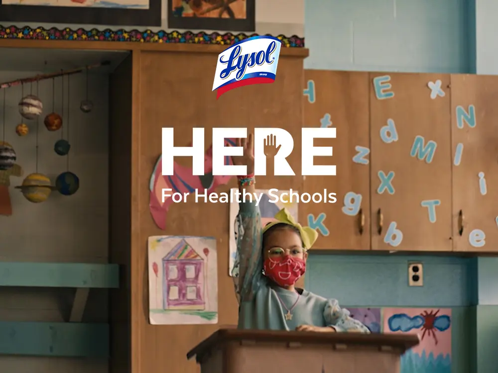 A student wearing a face mask in a classroom raises their hand to attract the attention of the teacher. The Lysol Here for Healthy Schools Logo overlays the image.