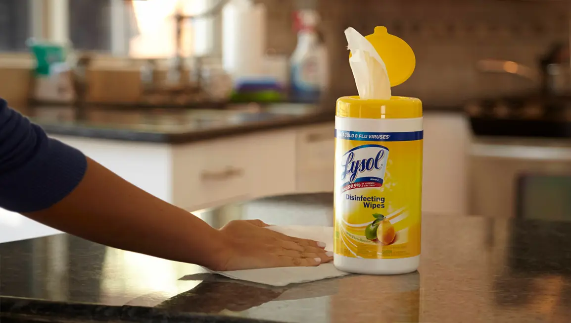 A canister of Lysol Disinfecting wipes on a kitchen counter. A hand is using a wipe to wipe down the counter next to the container.