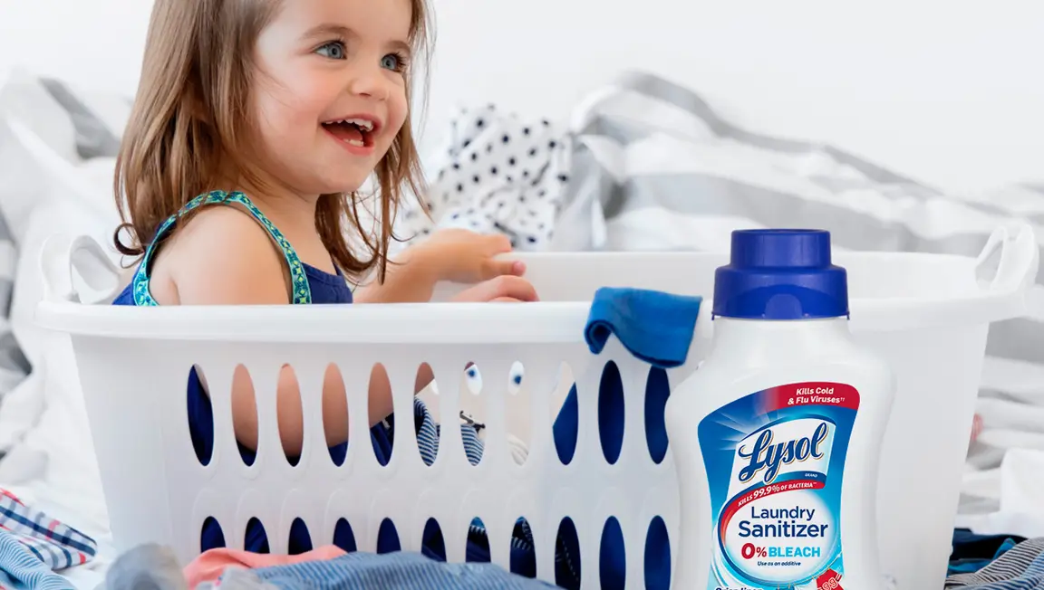 Young child sitting in plastic laundry basket and smiling surrounded by laundry and container of Lysol Laundry Sanitizer