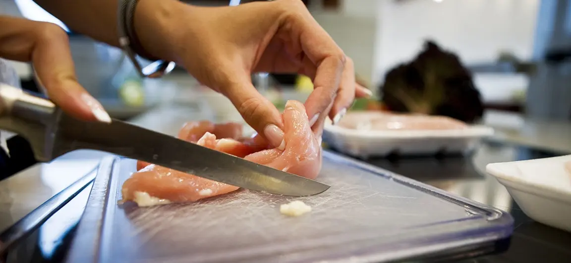 A sharp knife being used to cut up raw chicken on a plastic cutting board in a kitchen.