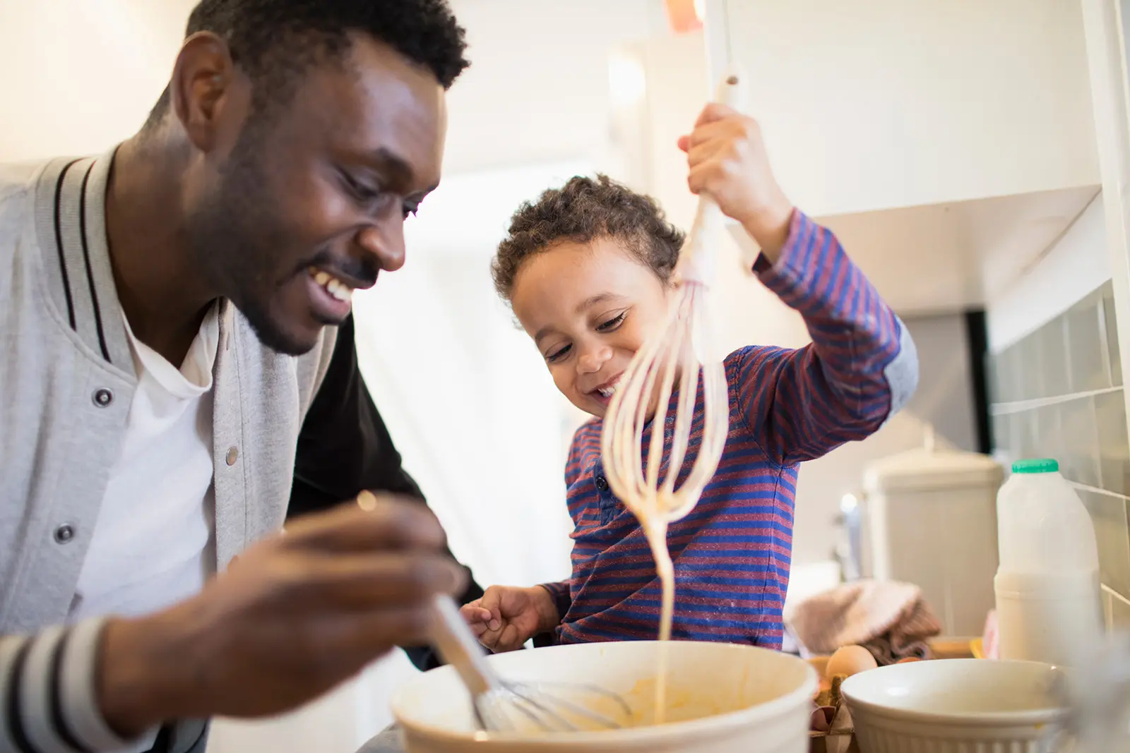 Parent stirring bowl of batter in kitchen next to child holding whisk dripping batter
