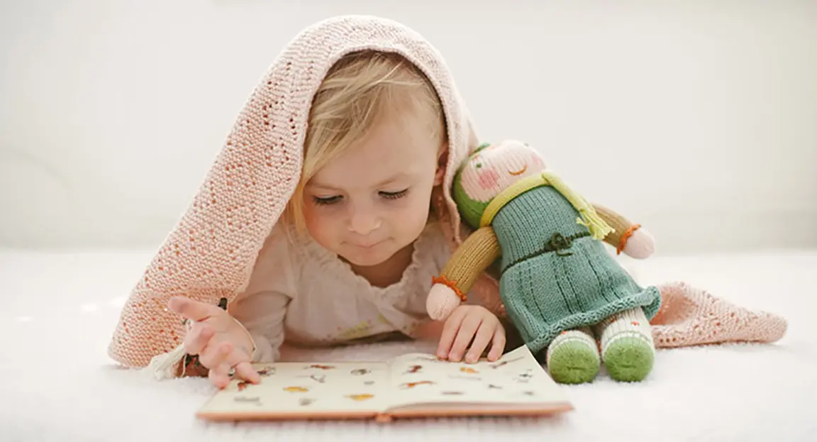 Young child with blanket draped on head reading book next to stuffed doll