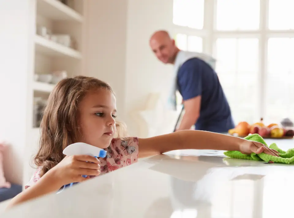 Child in foreground wipes kitchen counter with cloth and spray bottle while parent in background looks on