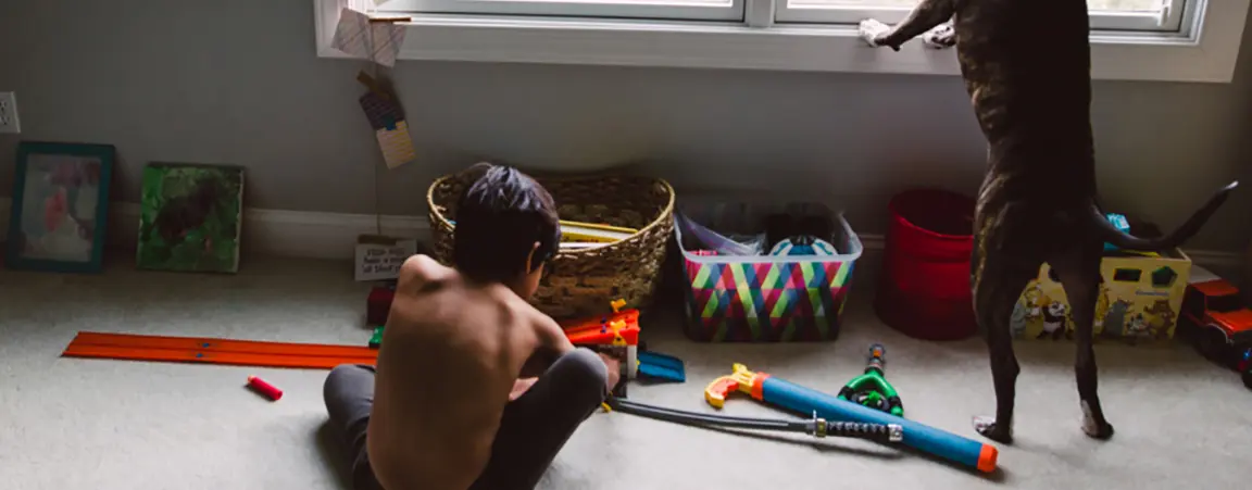 Child sits on floor surrounded by toys next to dog looking out window with front paws on windowsill