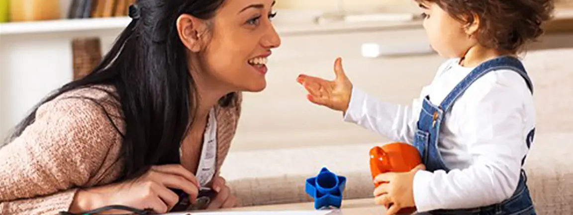 Smiling parent leaning towards toddler holding toy and child holds out hand towards parent