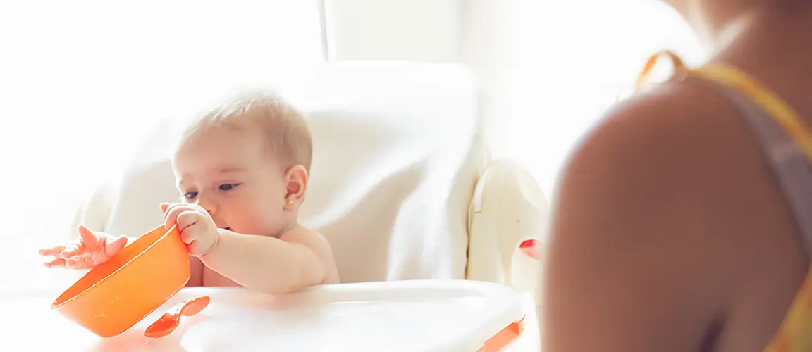 Baby in high chair tipping orange bowl while parent looks on