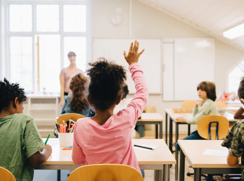 Child on second row of desks raises hand to alert teacher at front of classroom