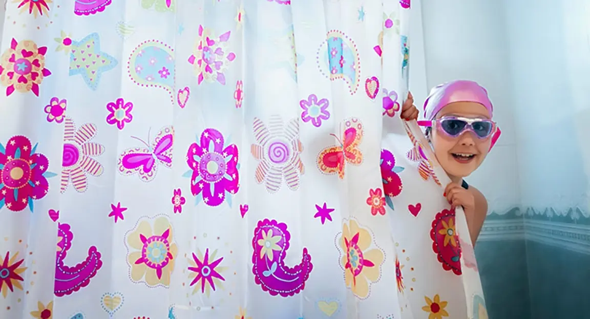 Child in shower cap and goggles peeking from behind floral shower curtain