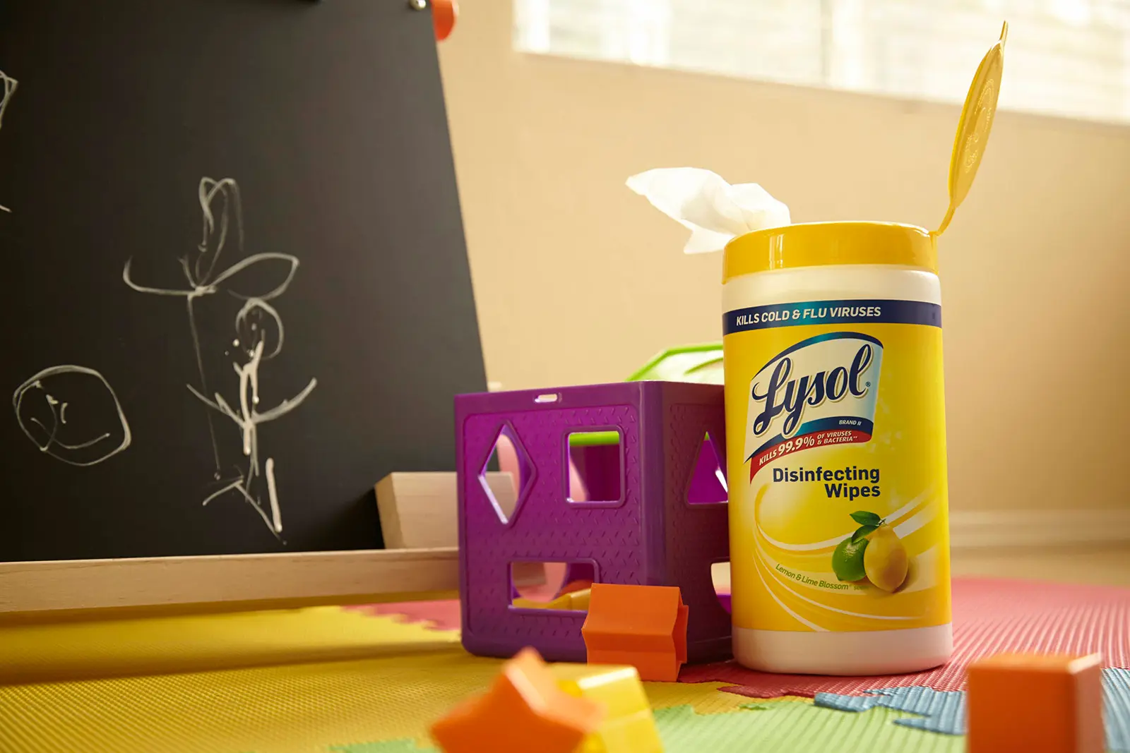 Canister of Lysol Disinfecting wipes on colorful foam floor next to toys and blackboard with chalk drawings