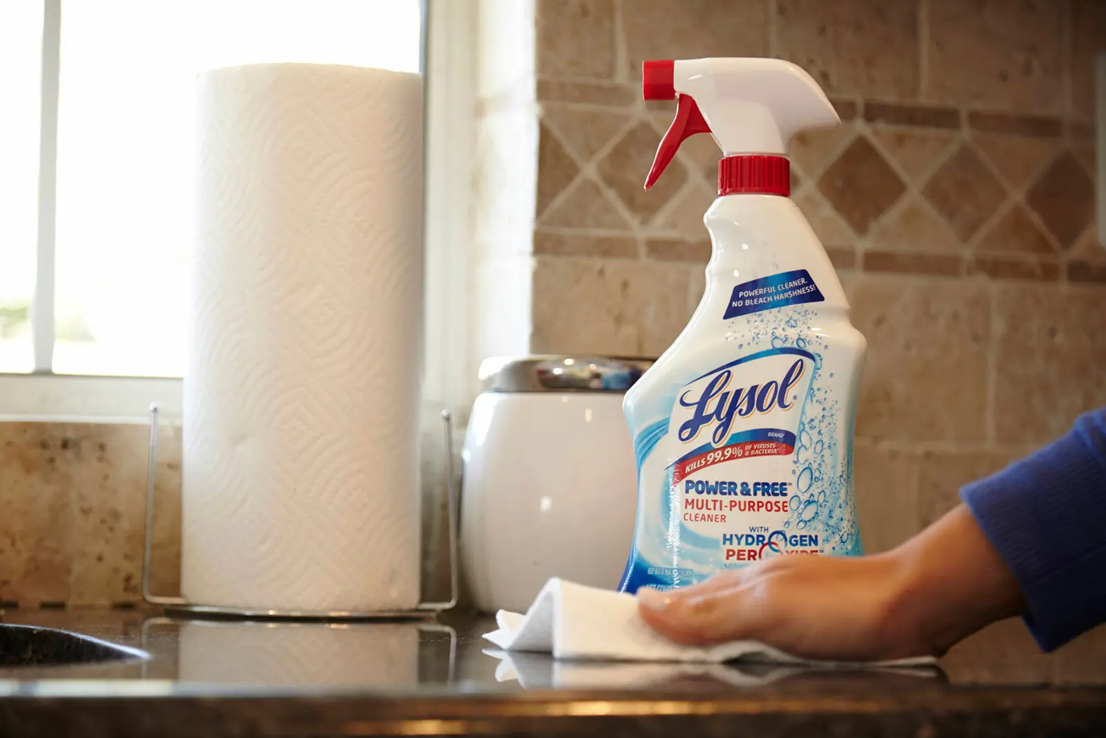 Person wiping a kitchen counter in front of a bottle of Lysol Multi Purpose Cleaner with Hydrogen Peroxide