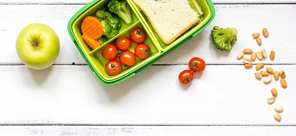A lunchbox containing healthy food sitting on wooden surface next to apple, tomatoes, nuts and broccoli