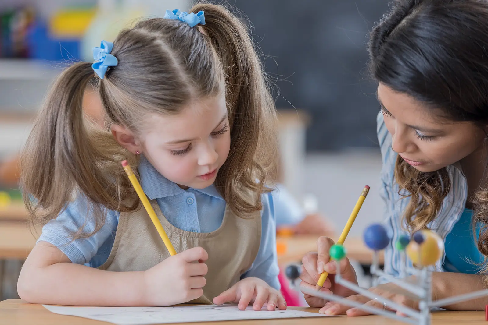 Teacher leans over child while both write on the paper in front of them