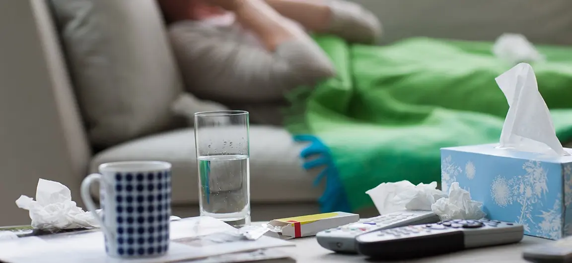 A sick person lying on a sofa and covered with a blanket. In the foreground discarded tissues rest on top of television remote controls.