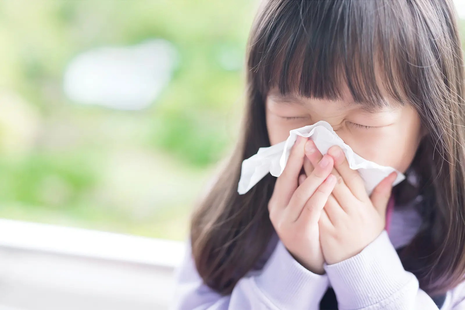 Child sneezing into tissue in front of window