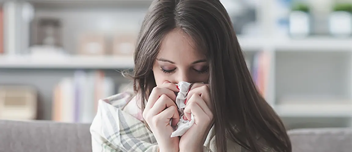 Adult wrapped in blanket on sofa holding tissue to their nose