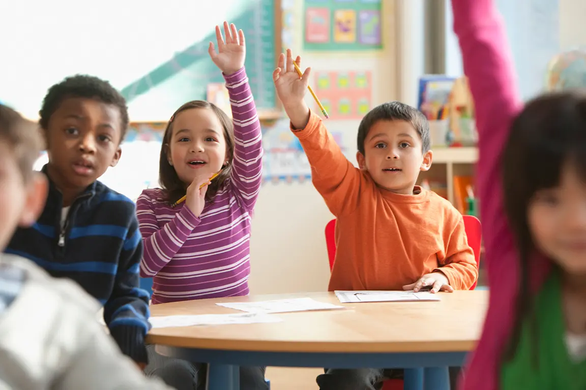 Children in classroom excitedly try to get the attention of the teacher