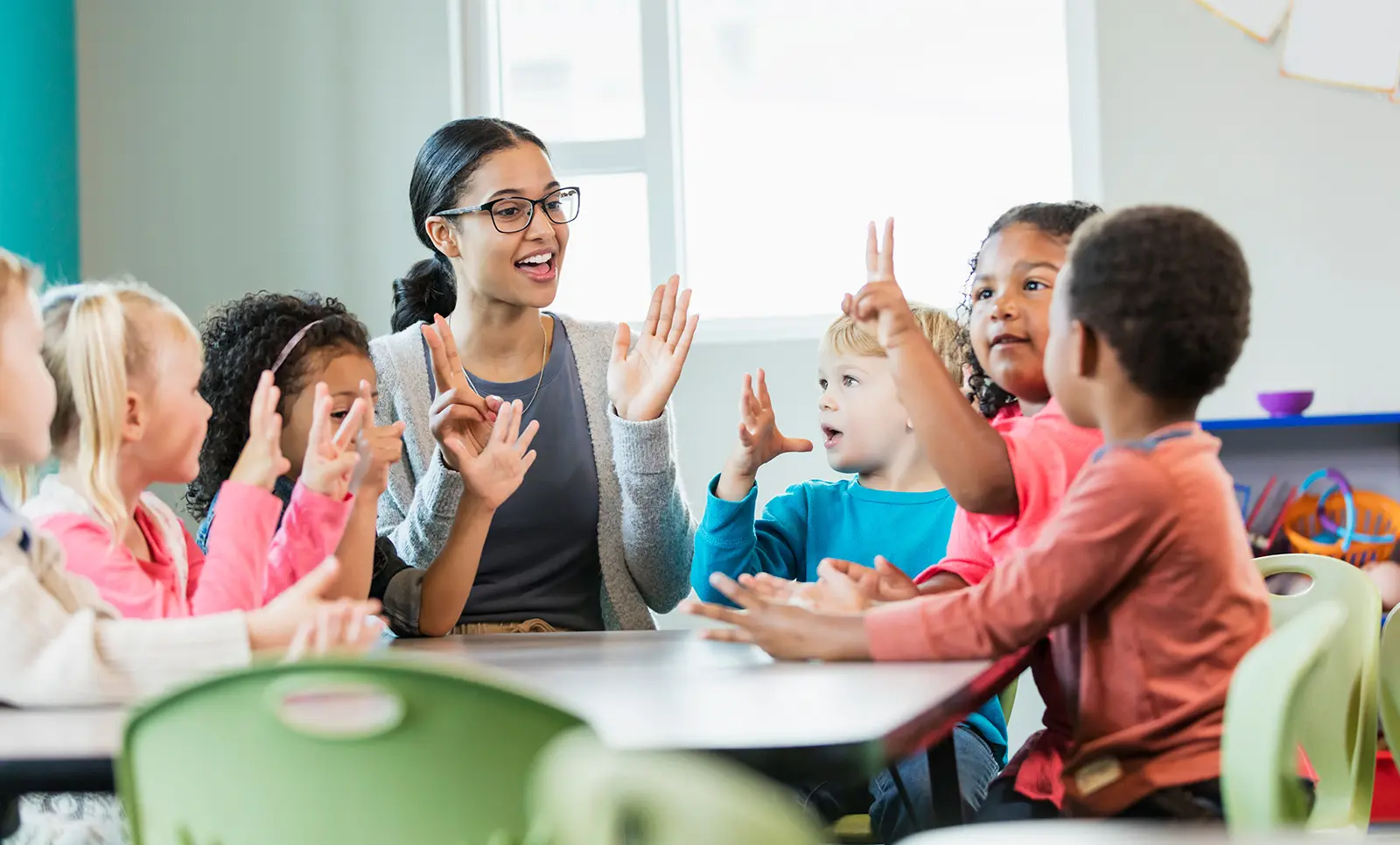 Teacher and six children sitting at school table talking with hands held out