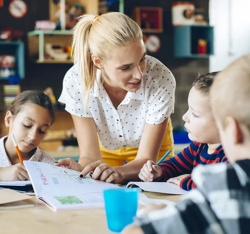 A teacher leaning over a desk and pointing to a textbook while their students look on.