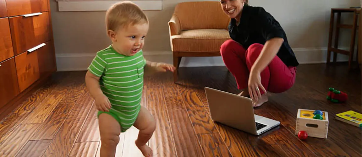 Parent crouching on floor behind laptop watches toddler walk nearby