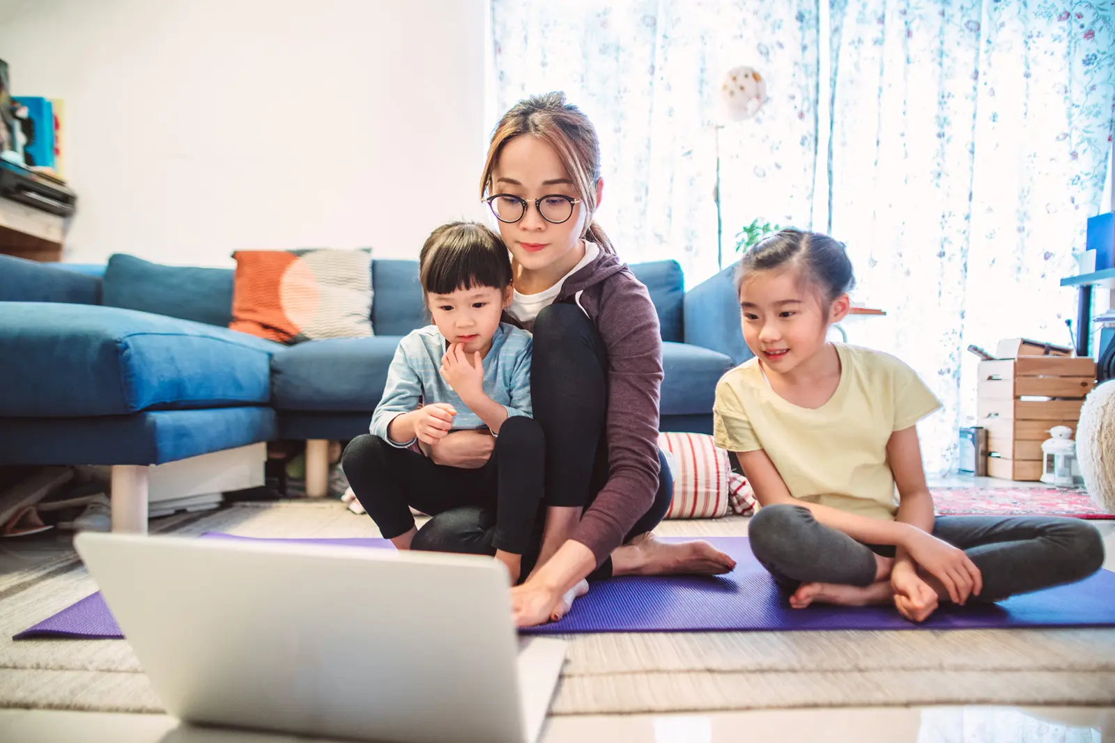 Parent with two children sitting on yoga mat in living room looking at laptop screen