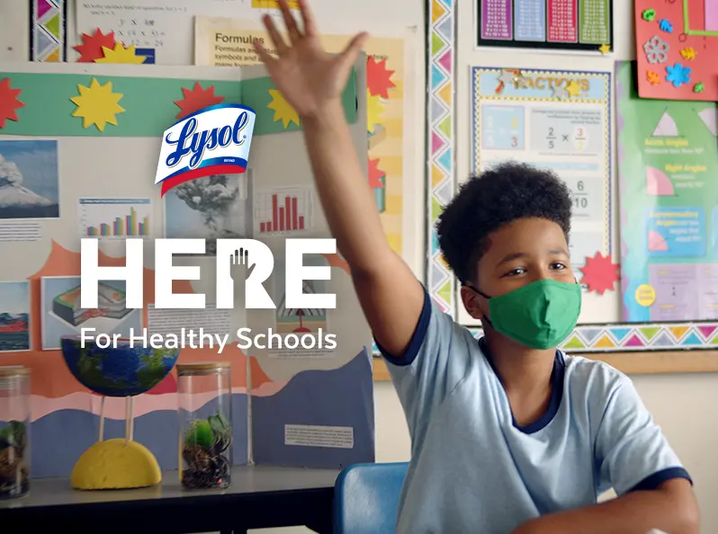 A student wearing a face mask raises their arm to attract the attention of a teacher. The Lysol Here for Healthy Schools logo overlays the image.