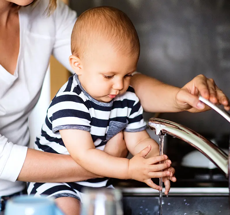 A parent holds their baby over the kitchen sink. The baby is holding their hands under the running faucet.