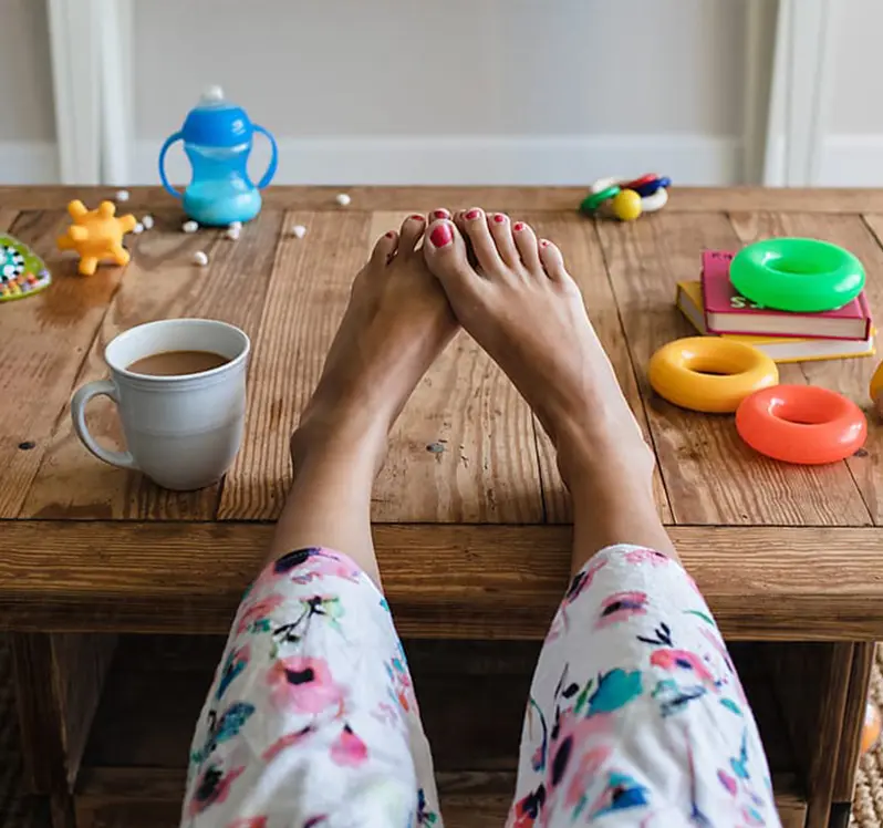Feet with painted toenails resting on a table with a coffee mug, children's toys and a sippy cup of milk.