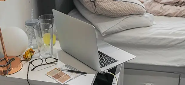 An open laptop sitting on a bedside table, next to a lamp, eyeglasses and empty drinking glasses