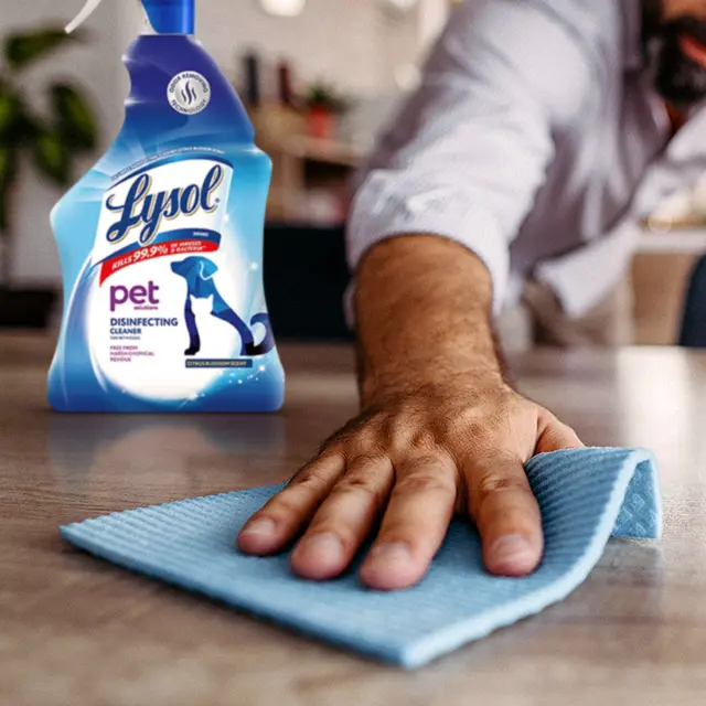 Male hand wiping table surface with a cloth, Lysol spray in background.
