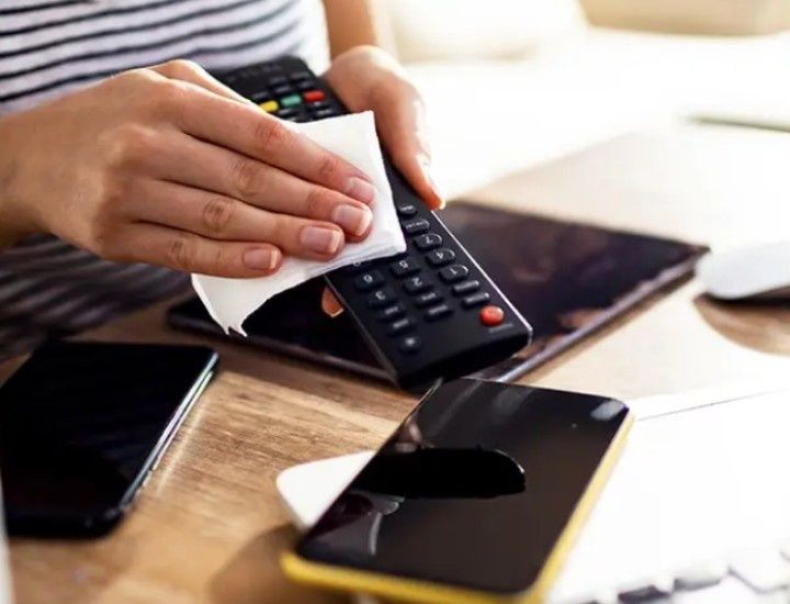 A person cleans a remote control with a cloth. Nearby, a smartphone with a black case and other electronics are on the wooden table.