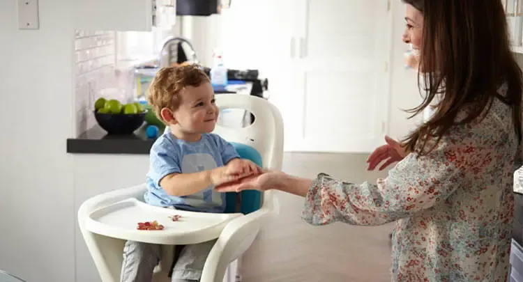 Toddler in high chair holding hand of parent stood next to it