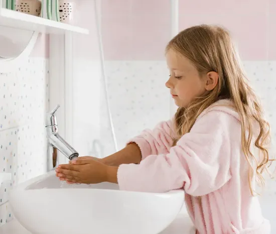 Young girl in a pink bathrobe washing her hands at a bathroom sink.
