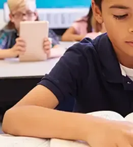 Boy reading a book in a classroom with other students behind him.