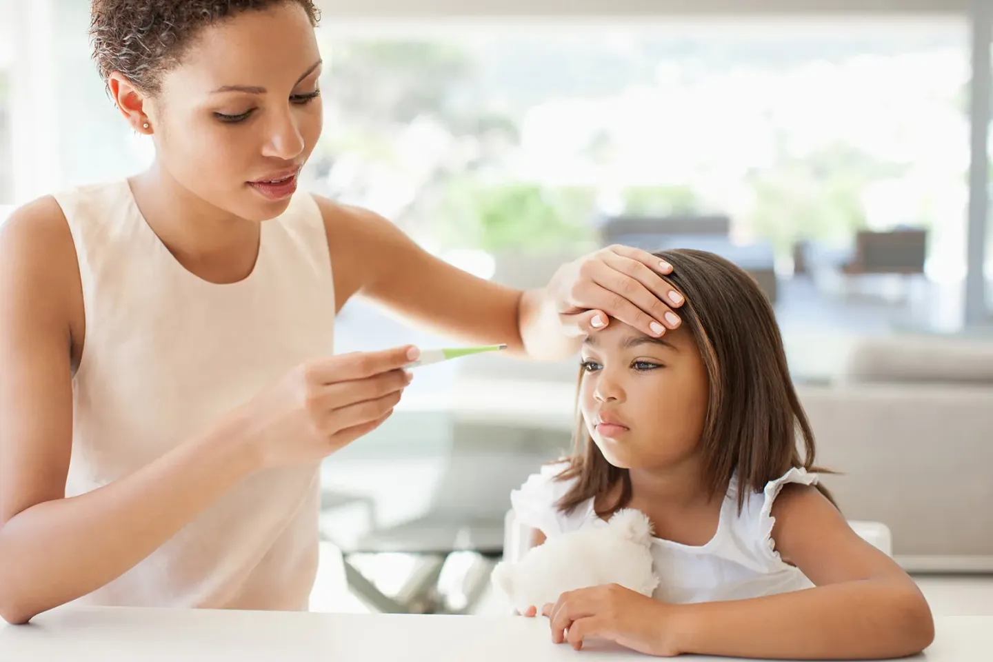 Parent looking at thermometer while holding hand to forehead of unhappy child