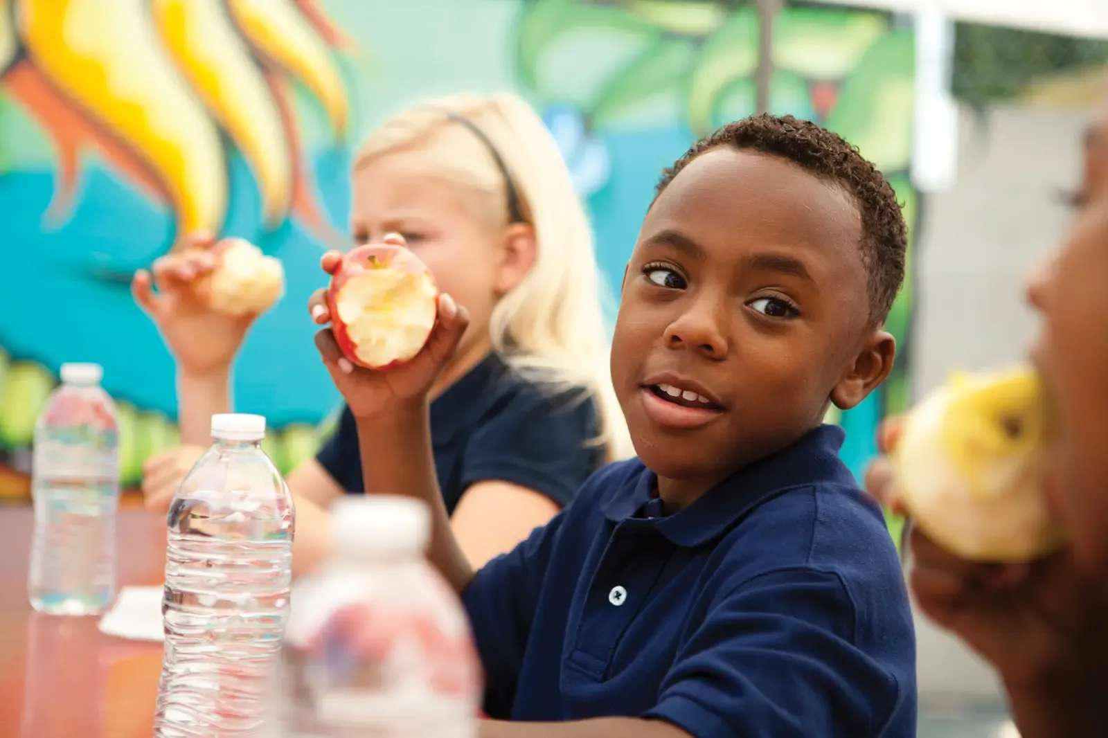 Children in school uniform sit at table eating apples