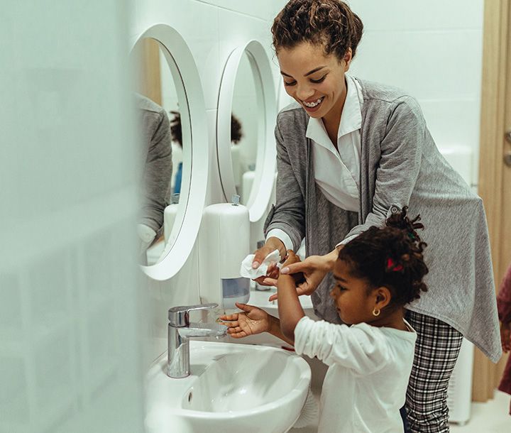 A woman helps a young girl wash her hands at a bathroom sink with a white oval mirror and a soap dispenser.