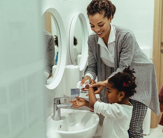 A woman helps a young girl wash her hands at a bathroom sink with a white oval mirror and a soap dispenser.