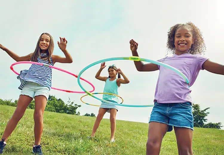 Three children stood in grass field spinning colorful hula hoops on their waists